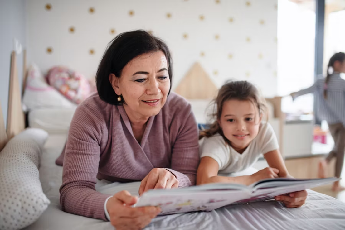 An upper middle-aged woman and a child smiling as they read a book together on a bed.